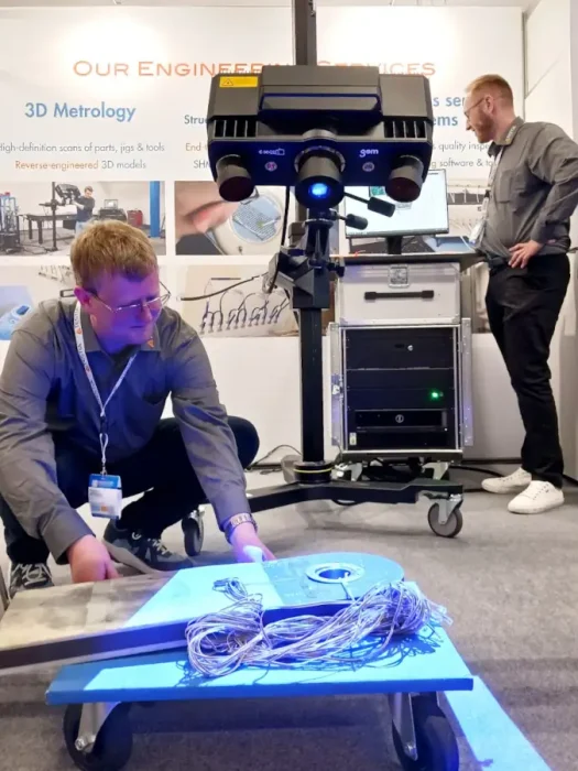 Two engineers are scanning an aircraft part using a GOM scanner. The scanner is a big device on a tripod that projects blue light onto the part and records the reflection with a stereo camera.